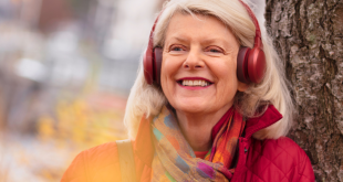 An older woman with grey hair smiling as she stands under a tree. She wears a red jacket, scarf and red headphones.